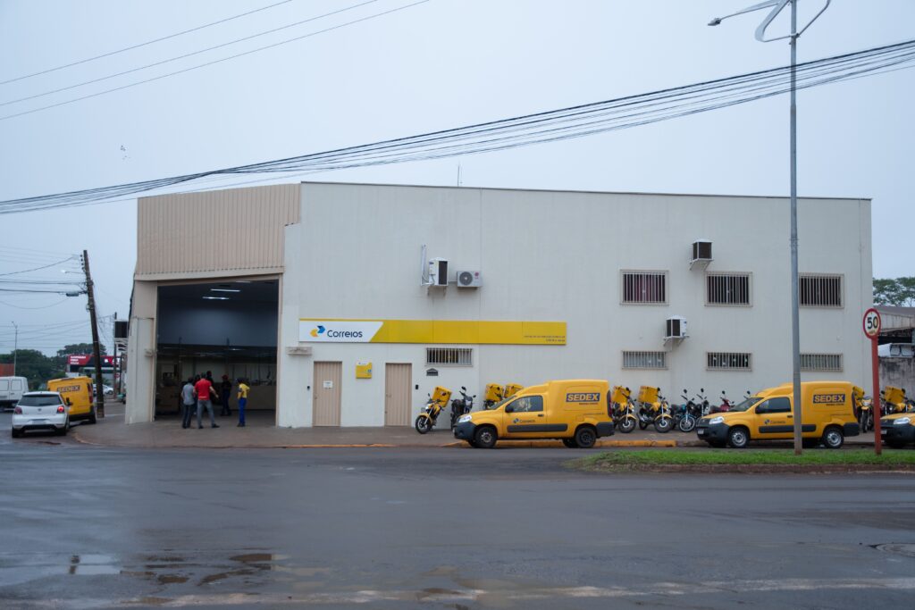 Imagem do CDD Rio Verde - prédio bege com fachada amarela onde está escrito Correios - carros e motos na cor amarela estacionados em frente ao prédio